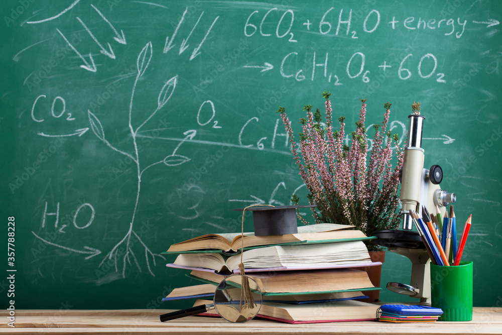 Education and sciences concept - books, graduation cap and microscope on the desk in the auditorium,