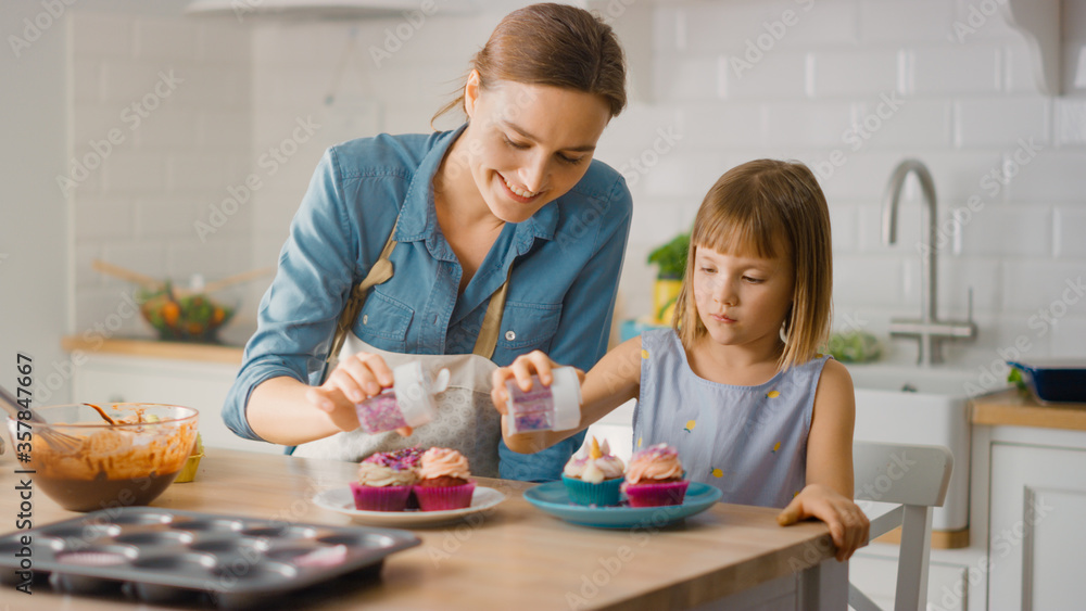 In the Kitchen: Mother and Cute Little Daughter Sprinkling Funfetti on Creamy Cupcakes Frosting. Fam