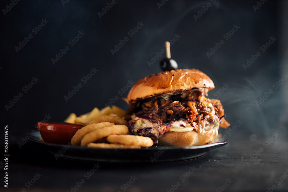 Juicy pulled pork burger on a plate with fries and onion rings. Dark background, copy space.