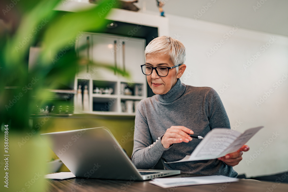 Senior woman doing finance at home, holding paper document and looking at laptop.