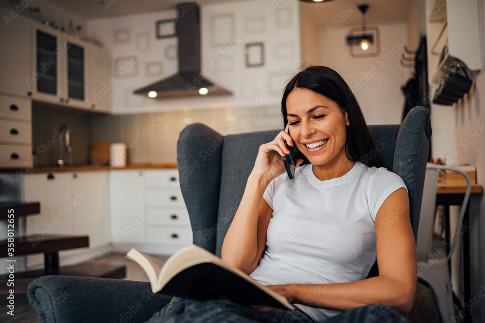 Portrait of a smiling woman reading book and talking on mobile phone.