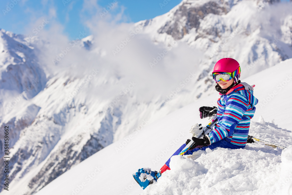 戴着粉色头盔和滑雪装备的漂亮女孩坐在雪地里的山顶上观察比赛