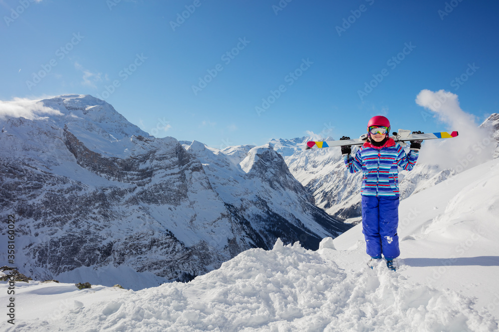 Happy smiling girl in colorful outfit, pink helmet and color glasses hold ski on her shoulders stand