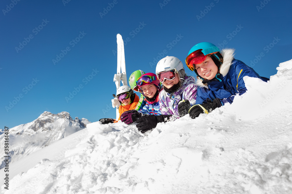 Big group of ski kids lay on snow cheerfully smiling over mountain peak and blue sky in colorful spo