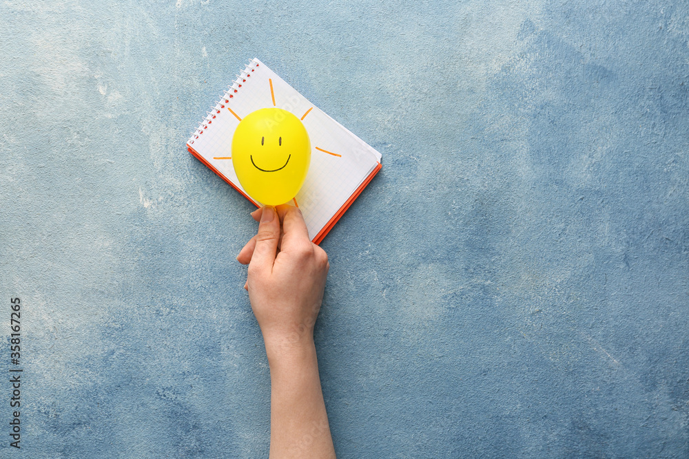 Female hand holding air balloon with drawn happy face and notebook on color background