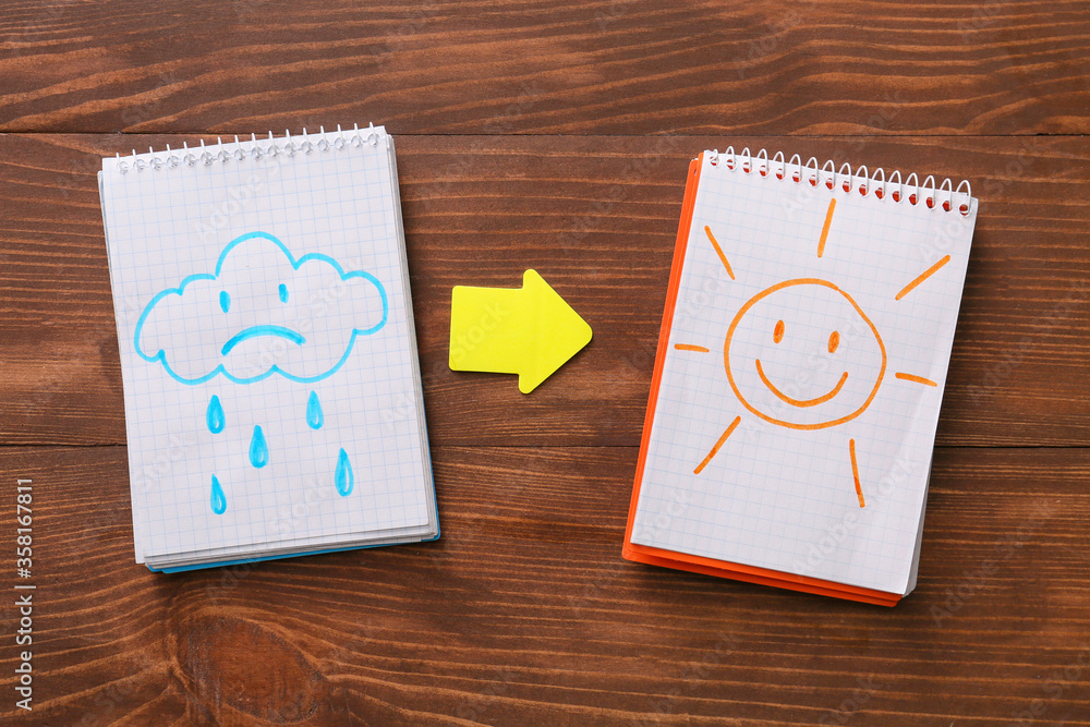 Notebooks with drawn sad cloud and happy sun on wooden table