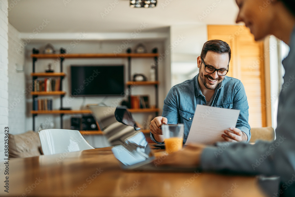 Smiling man reading a letter at home, sitting at the table and his wife using laptop.