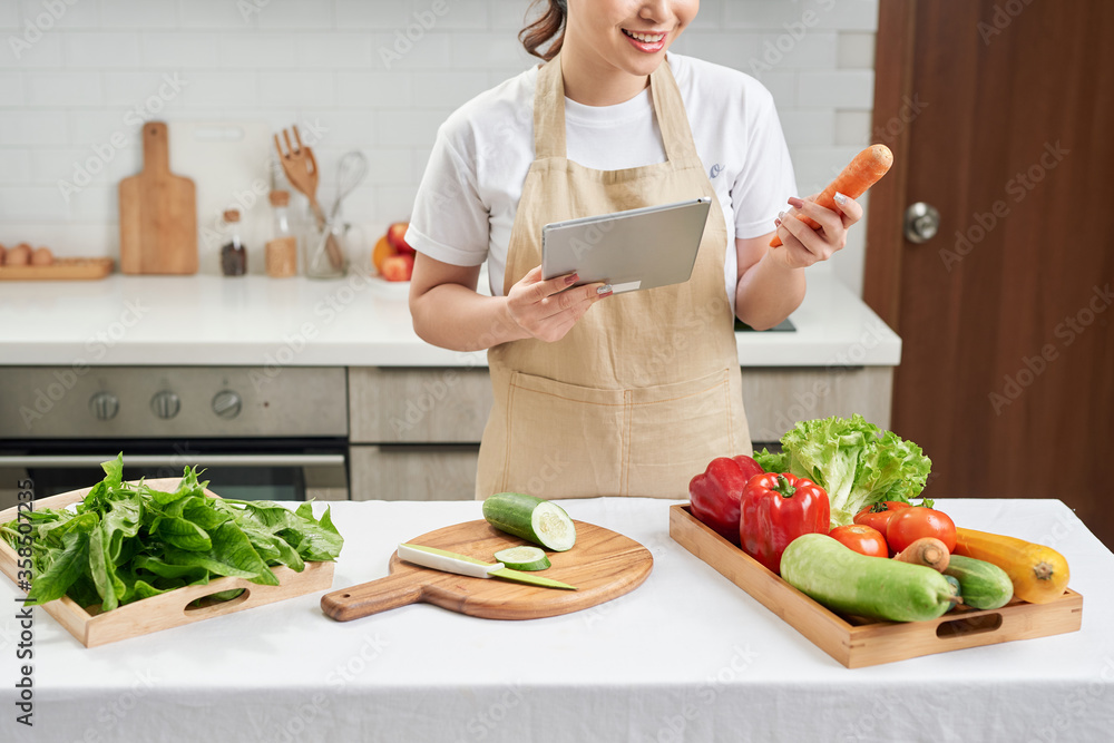 beautiful korean woman in apron holding tablet pad looking at list and checking fresh vegetables.