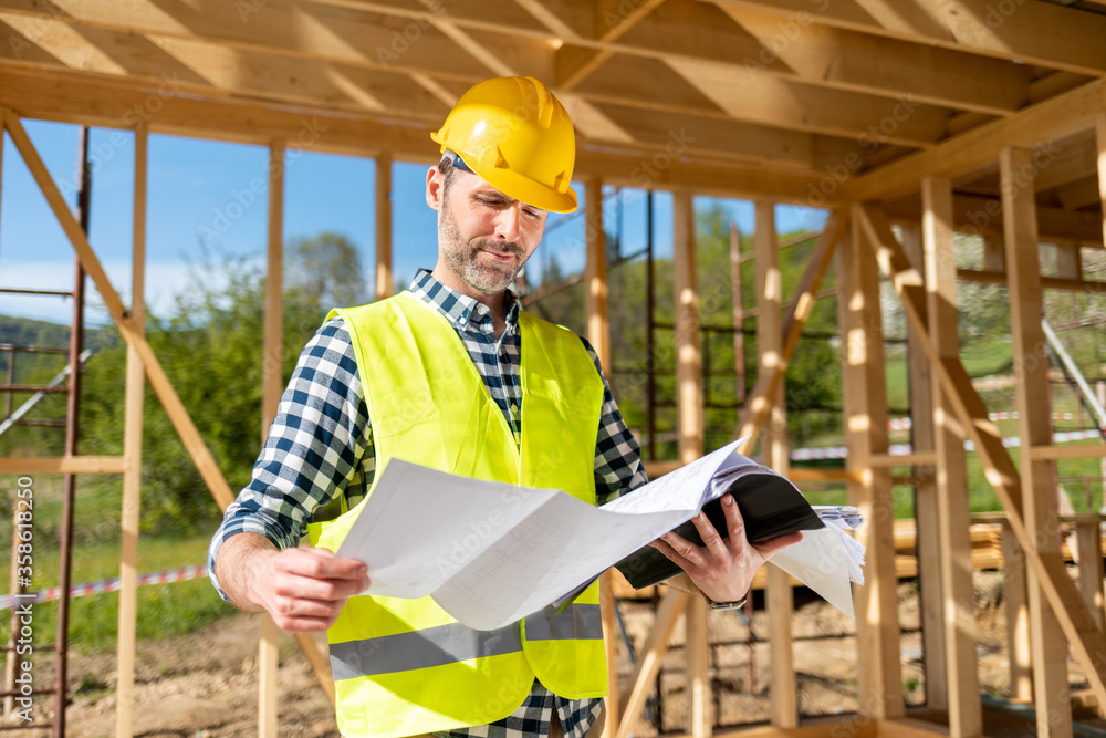 Engineer with hardhat and blueprints on building site of wood frame house under construction