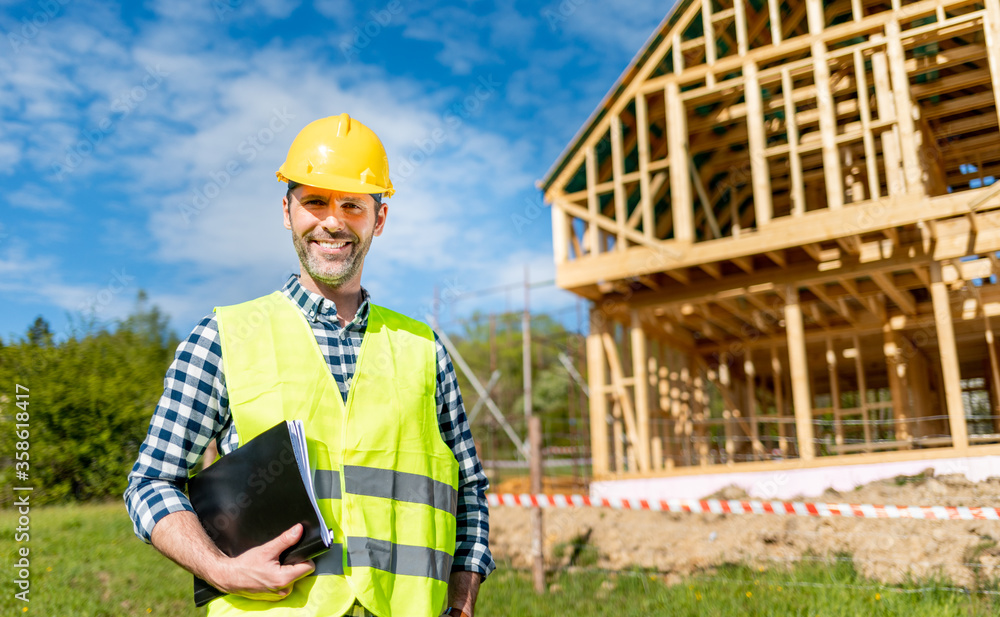 Engineer with hardhat and blueprints on building site of wood frame house under construction