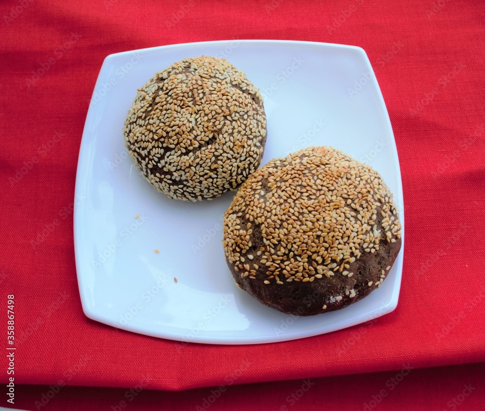 Homemade rye buns with sesame seeds on white plate, on the red linen fabric background