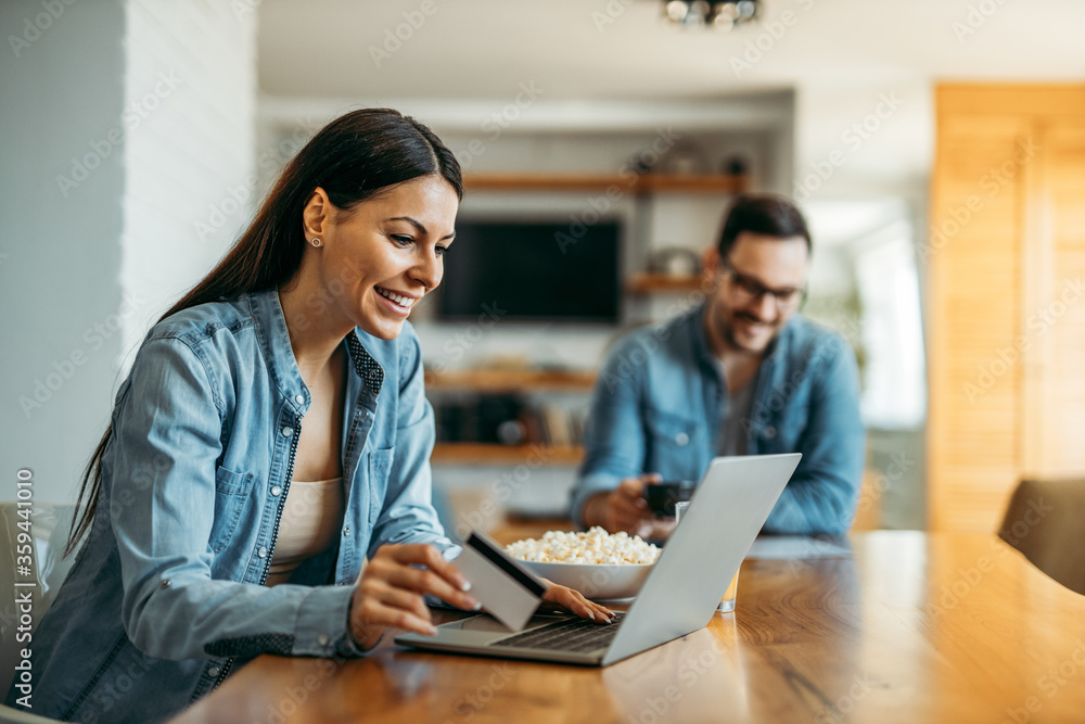 Relaxing at home. Portrait of a beautiful woman shopping online with credit card at home, husband in