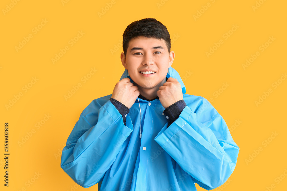 Young Asian man in raincoat on color background