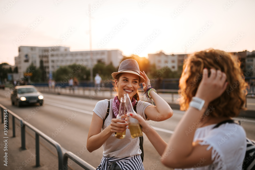 在阳光明媚的城市街道上，两个漂亮的时髦女人拿着装在瓶子里的饮料欢呼。