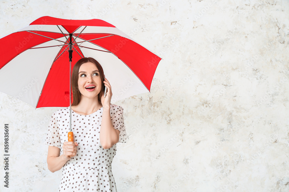 Beautiful woman with umbrella talking by mobile phone on light background