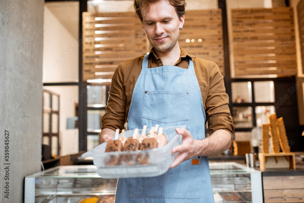 Portrait of a cheerful salesman in apron holding a tray full of chocolate ice cream on a stick while