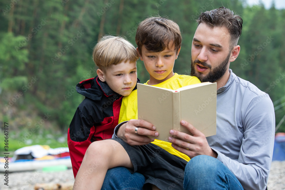 A man and two children (boys) reading a book together in the fresh air, in the background a lake