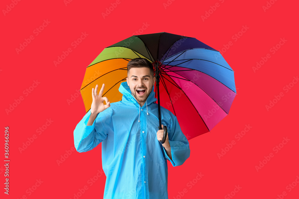 Young man in raincoat and with umbrella showing OK on color background