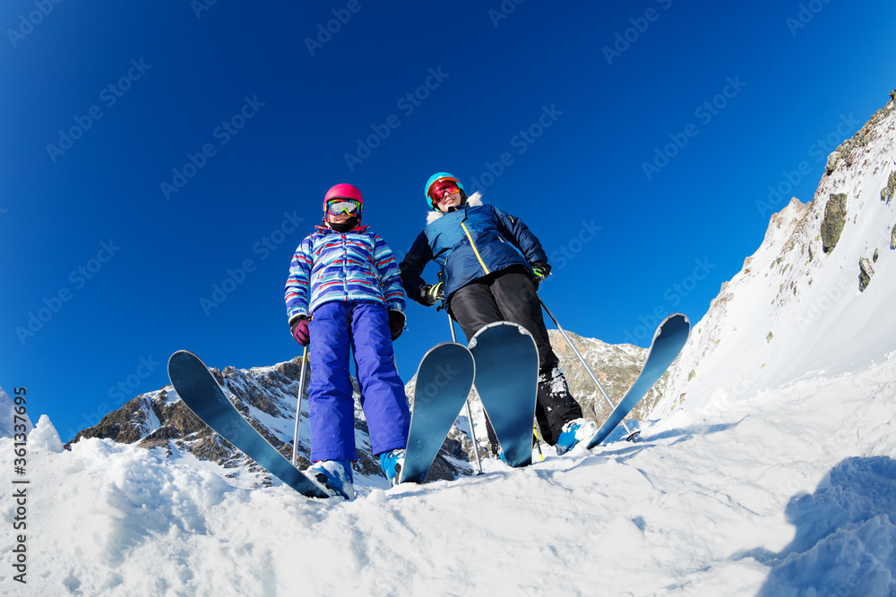 View from below of two girls stand on ski wearing helmets and masks on top of the mountain