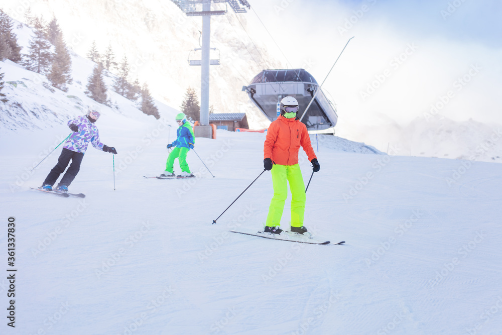 Three children ski downhill together in colorful outfit on Alpine mountain slope with lift station o