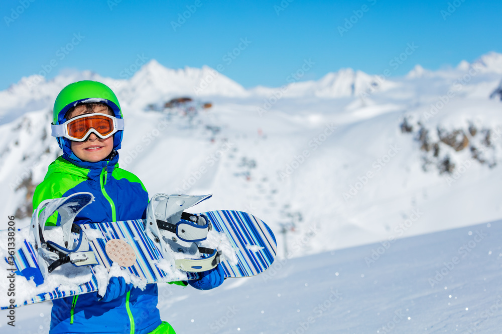 可爱的小男孩拿着滑雪板，背景是山地雪冬季度假村和滑雪缆车