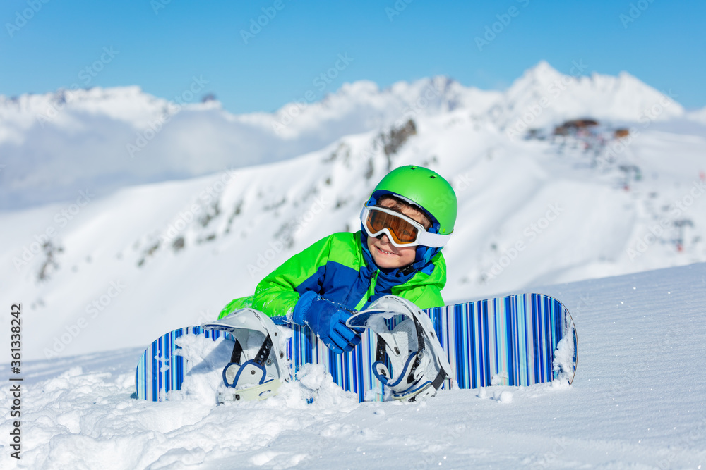 阳光明媚的度假山顶上，一个可爱的小男孩拿着滑雪板躺在雪地里的肖像