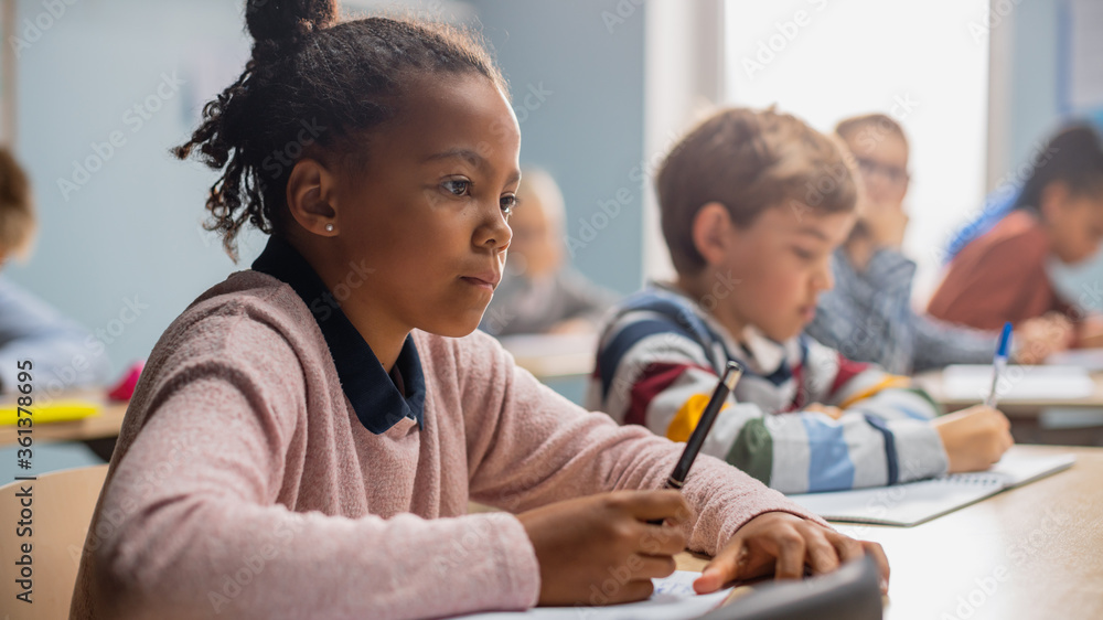 In Elementary School Classroom Brilliant Black Girl Writes in Exercise Notebook, Taking Test and Wri