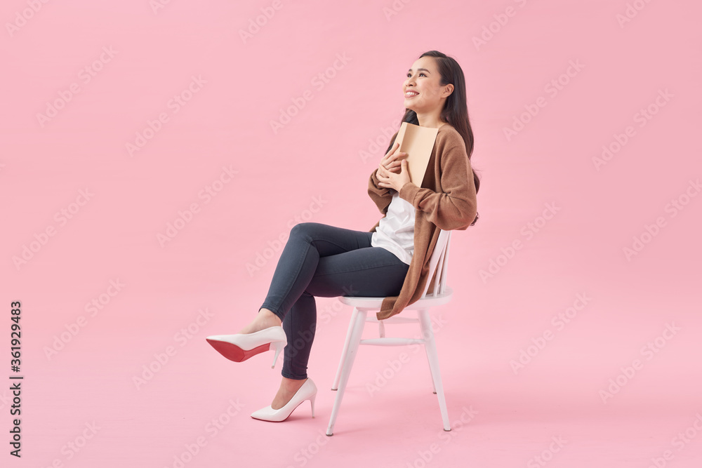 Young woman with book sitting on chair over pink