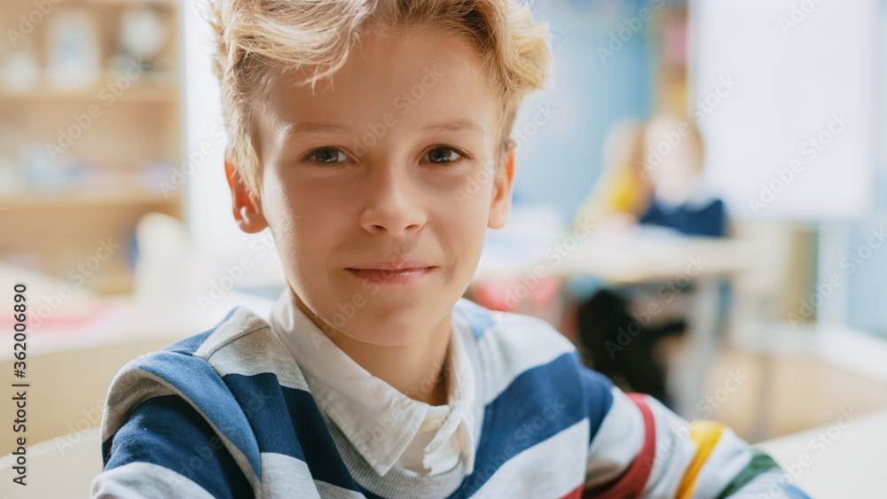 Portrait of a Cute Little Boy with Wavy Blond Hair Sitting at his School Desk, Smiles Happily. Smart