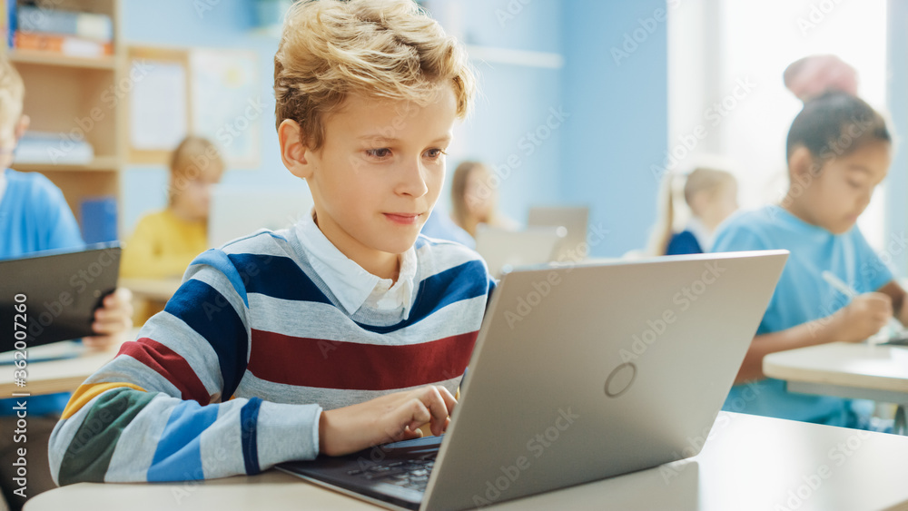 Elementary School Computer Science Class: Smart Boy Uses Laptop Computer, His Classmates work with L