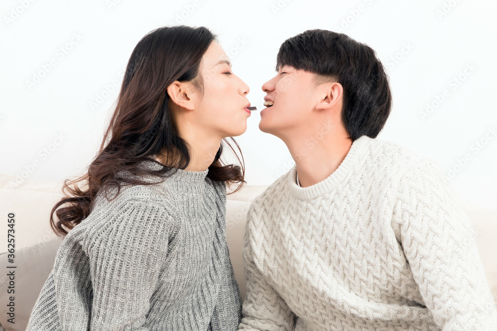 In winter, a pair of happy young Asian lovers sit on the sofa, mouth to mouth with each other, eatin