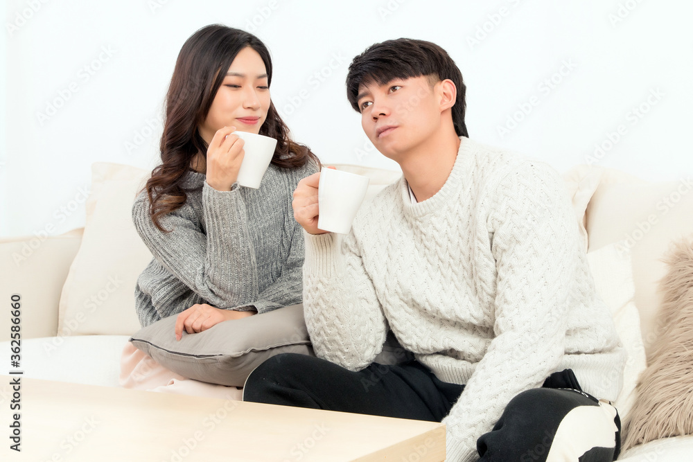 In winter, a pair of intimate young Asian lovers sit on the sofa, happily drinking hot coffee

