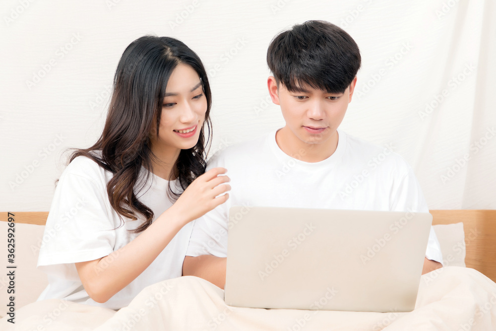 A pair of young Asian lovers sitting on the bed in white short sleeves are happily playing with comp