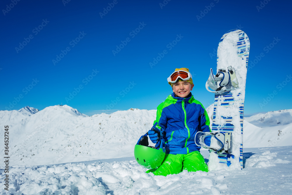 小男孩带着滑雪板坐在山脉上空的雪地里，戴着头盔，戴着滑雪谷歌眼镜