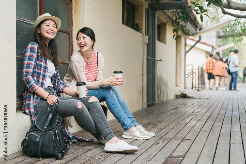 两位亚裔中国女性朋友坐在窗台上，拿着纸杯在户外喝咖啡