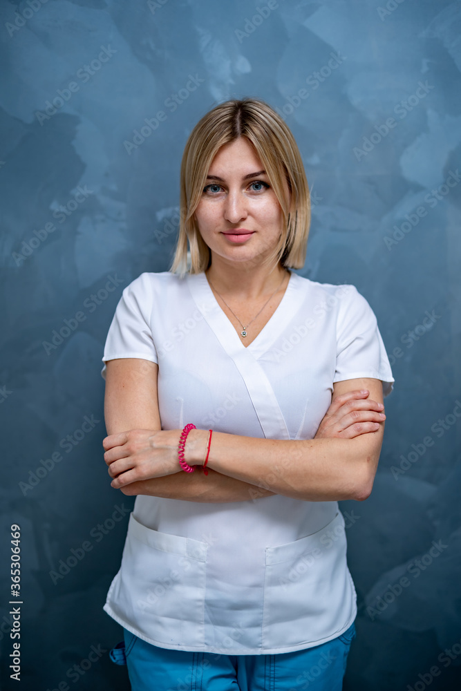 Waist portrait of female doctor wearing white scrubs and looking at camera posing against blue wall.