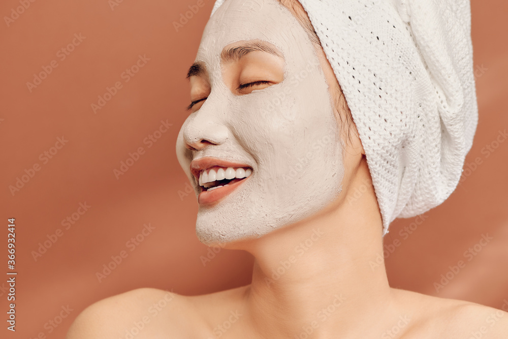 Close-up portrait of attractive girl with a towel on head and clay mask on face isolated over orange