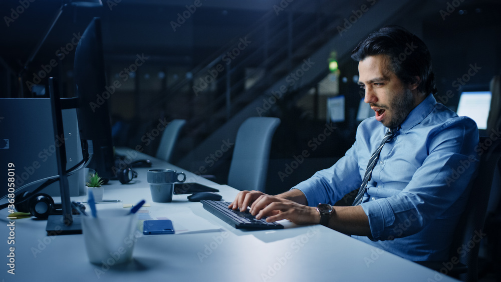 In the Office at Night Overworked Tired Office Worker Uses Desktop Computer Yawning, Drinks Coffee. 