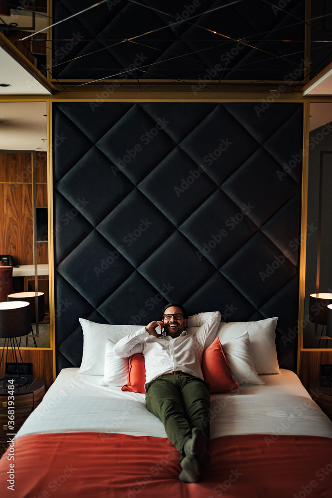 Smiling businessman relaxing at the hotel room. Lying on the bed and talking on smart phone, portrai