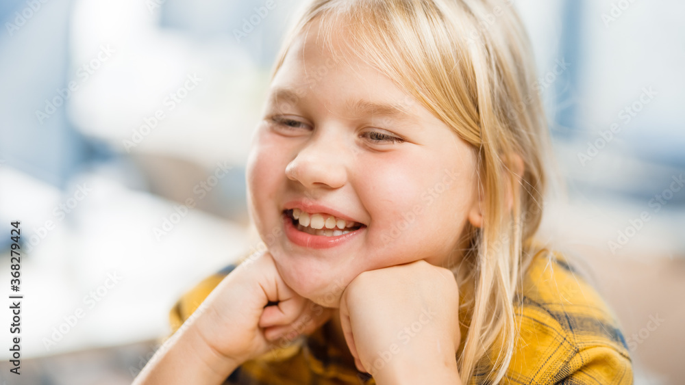 Portrait of a Cute Little Girl with Blond Hair Sitting at her School Desk, Smiles Happily, Shows Tee