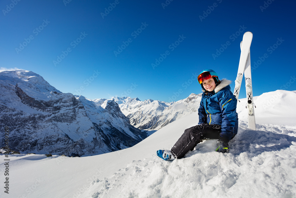 一个戴着蓝色头盔、穿着滑雪服、摘着口罩的女孩坐在山上的画像