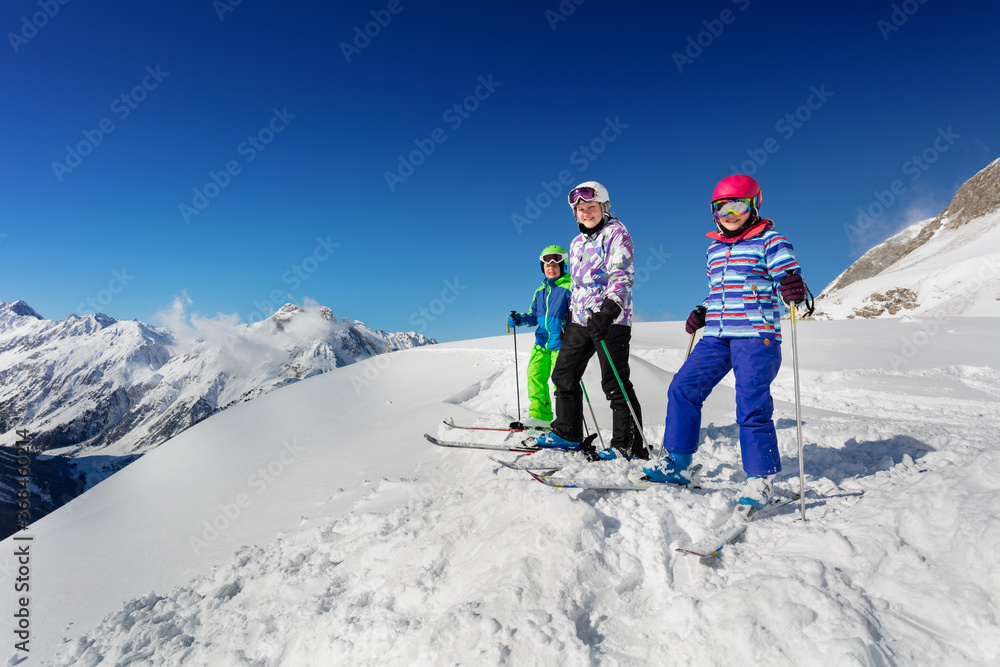 三个孩子一组站在山顶上，穿着五颜六色的滑雪服，在蓝天上