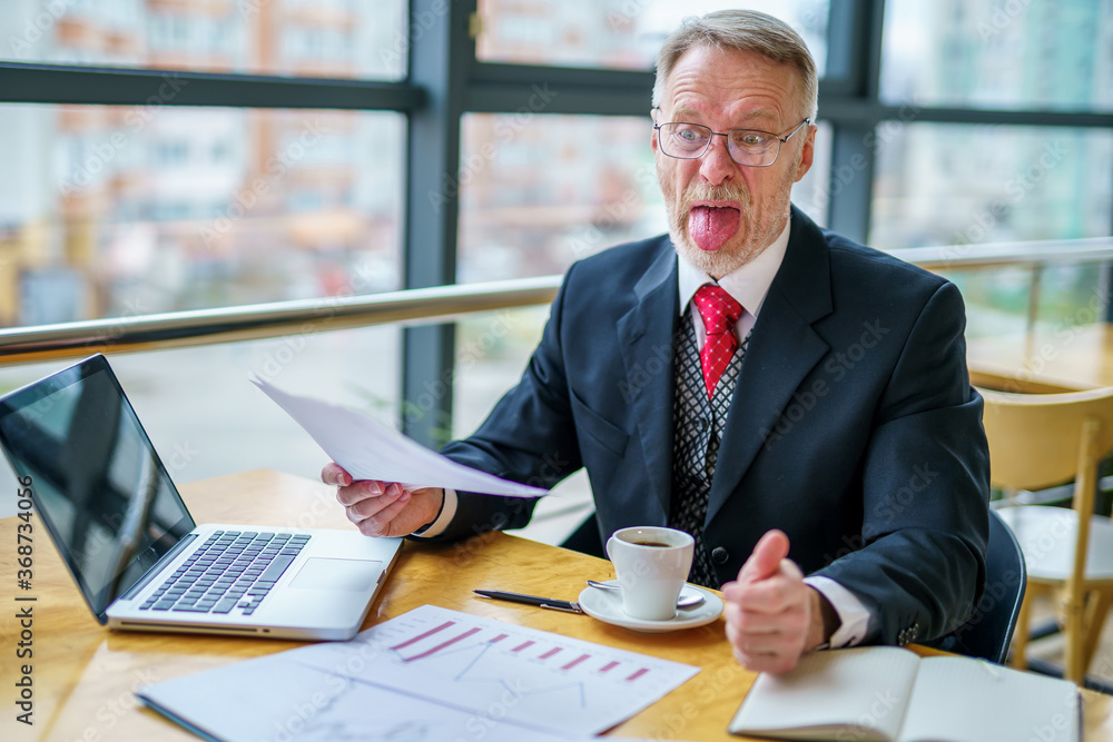 Happy mature business man shows tongue. Successful senior businessman sitting in meeting room with d