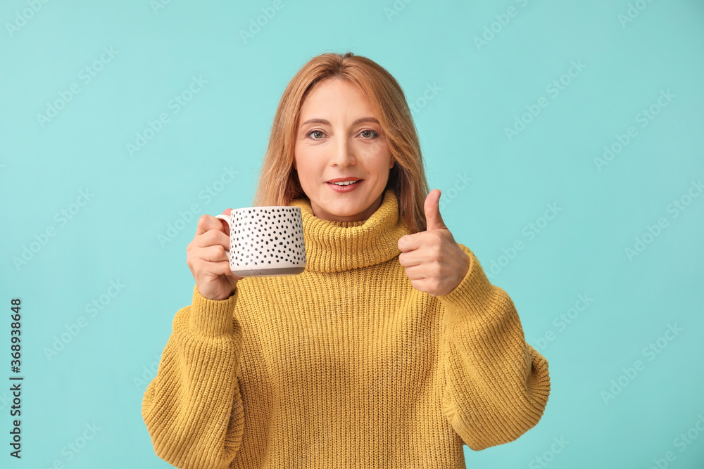 Beautiful woman with cup of tea showing thumb-up gesture on color background