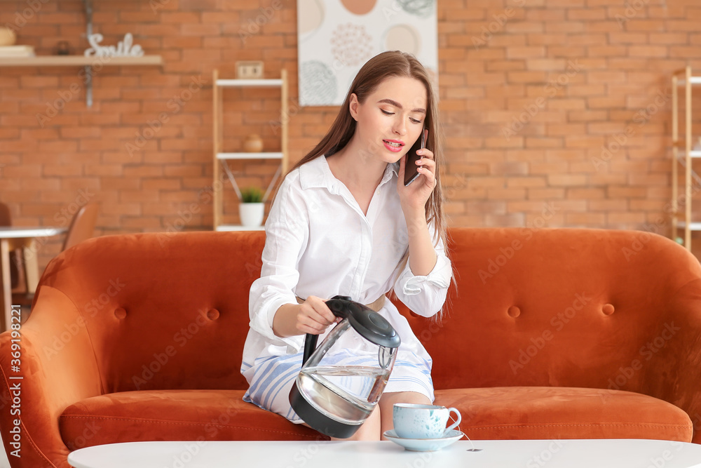 一个拿着手机的女人把电水壶里的热开水倒进家里的杯子里