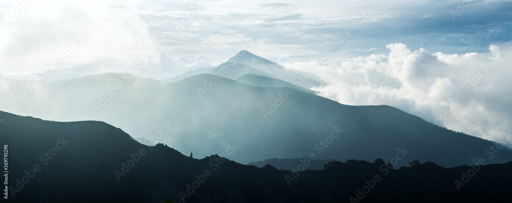 美丽的全景——蓝色雾蒙蒙的山脉。多云的天空和散发着光芒的薄雾的黑暗山峰。