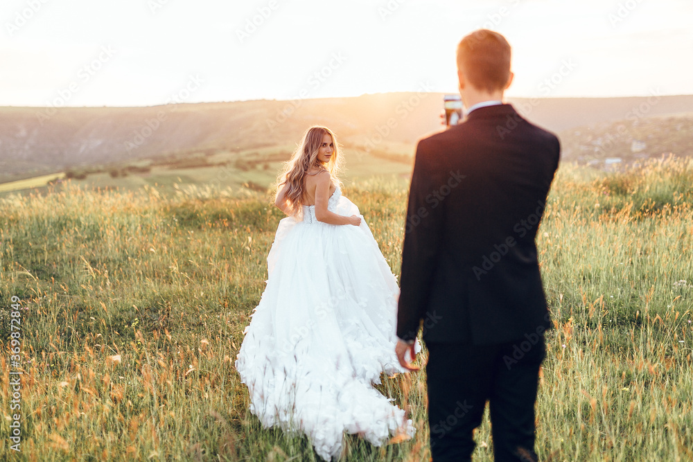 Bride and groom walking in the field. Groom taking a photo of his wife on his smartphone. Happy wedd