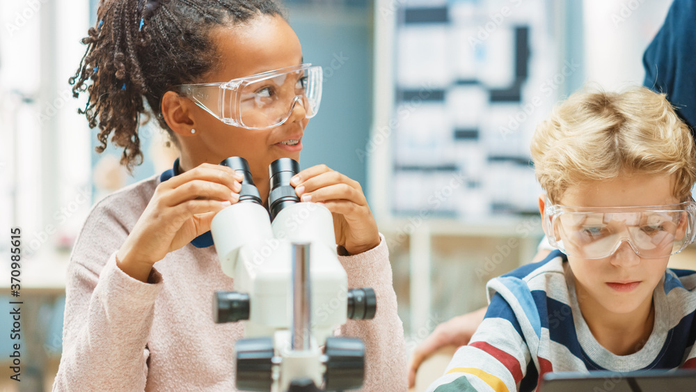 Elementary School Science Classroom: Cute Little Girl Looks Under Microscope, Boy Uses Digital Table