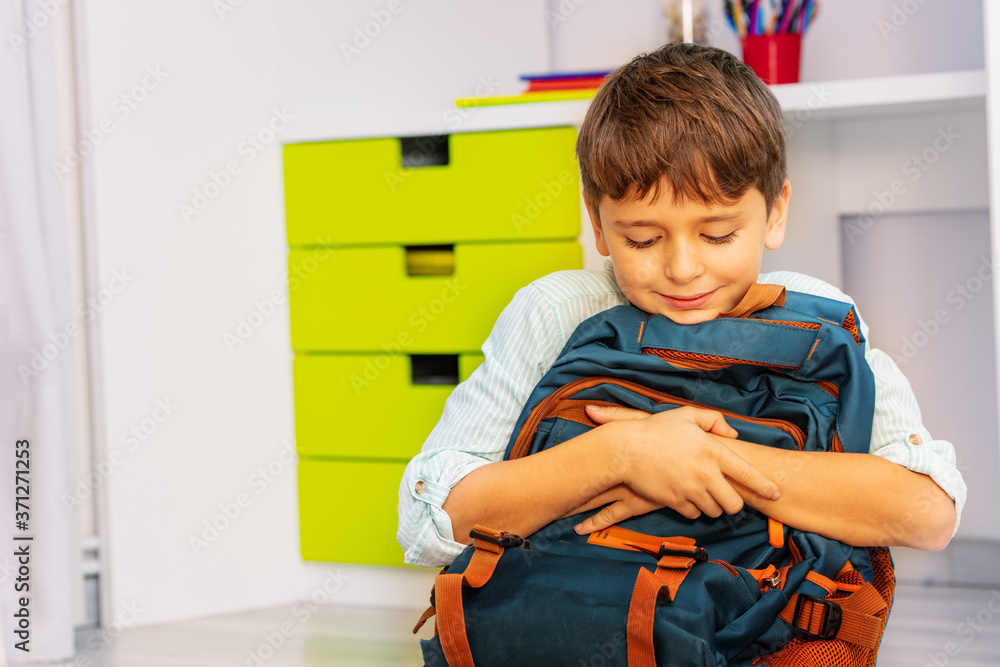 Close portrait of a smiling calm little boy sit in his room with positive expression holding school 