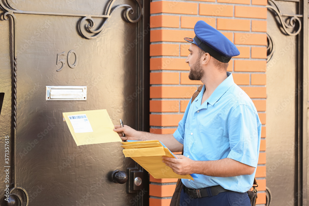 Handsome young postman putting letters in mail box outdoors